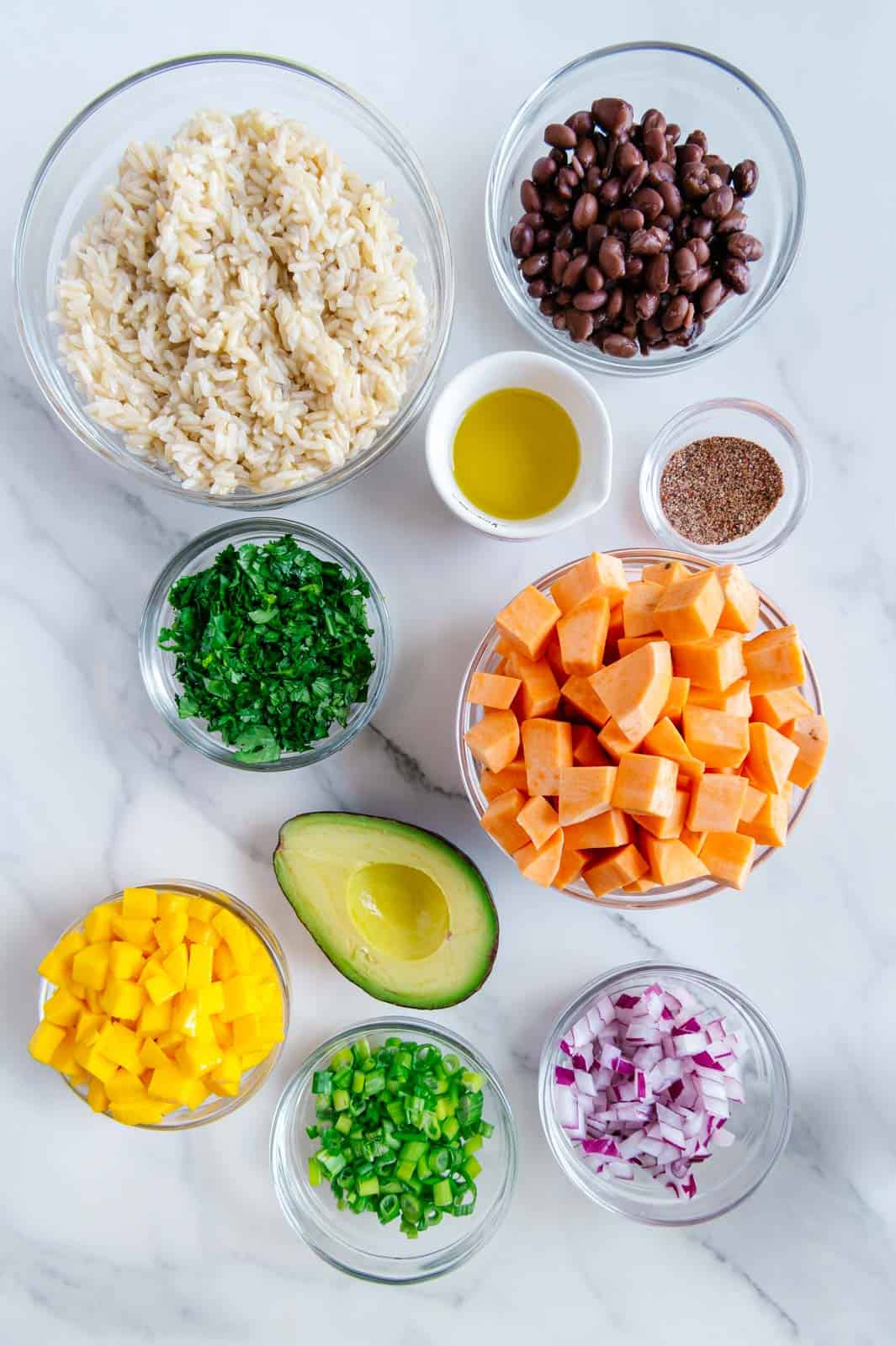 Bowls of brown rice, black beans, taco seasoning, fresh cilantro, diced sweet potatoes, red onion, green onion and diced mangos next to a half of an avocado with a marble white background.