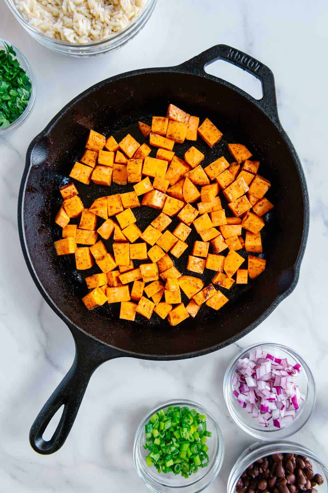 Raw sweet potatoes in a cast iron skillet next to diced red onion, diced green onion and black beans.