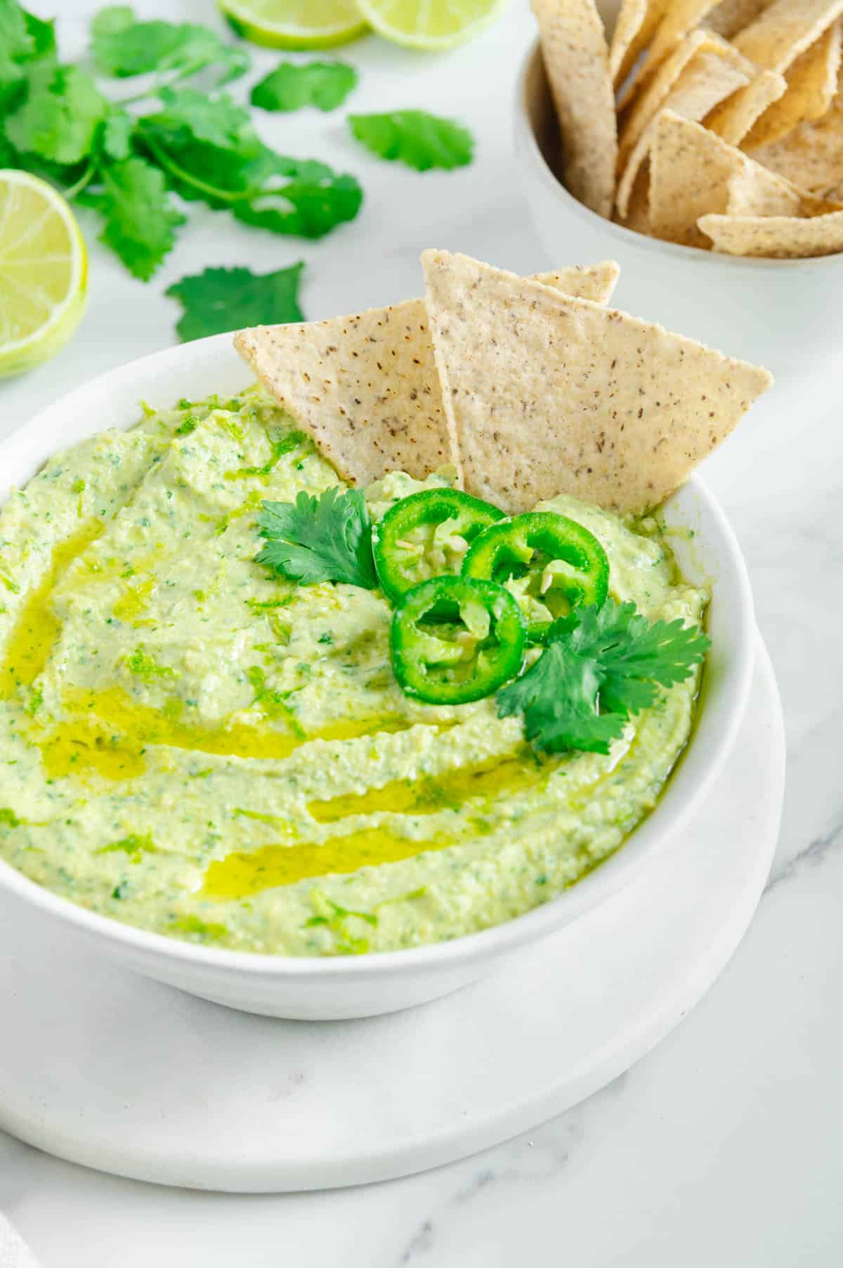 cilantro jalapeno hummus topped with olive oil, cilantro and jalapeno slices with tortilla chips dipped inside.