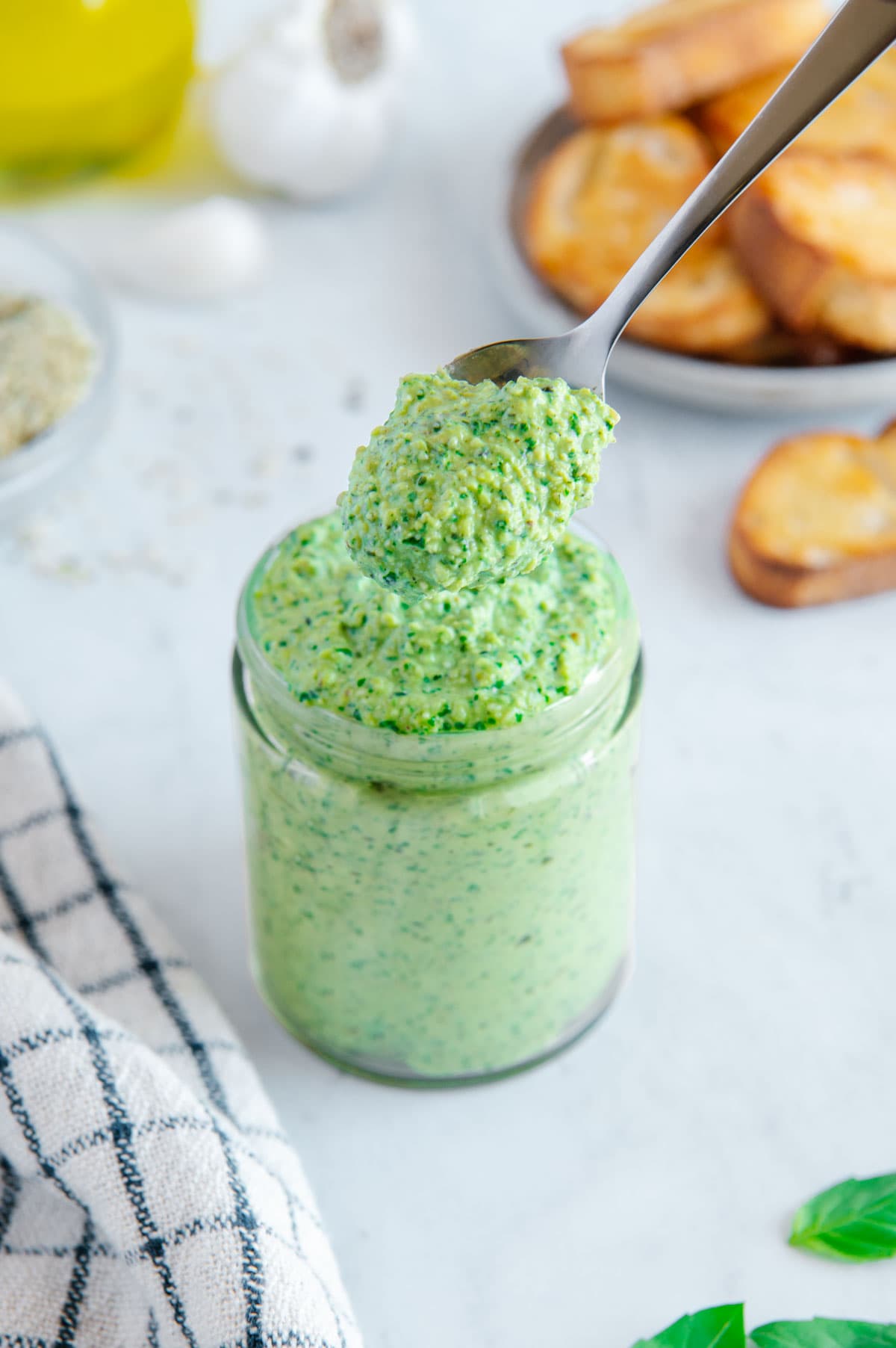 Vegan Hemp Heart Pesto in a glass jar with a silver spoon scooping some out next to a checkered white and blue towel with out of focus slices of toasted garlic bread behind it.