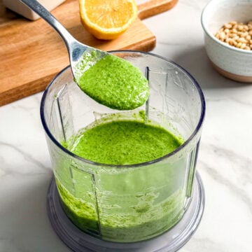 A spoon holds up vibrant vegan pesto above a food processor bowl of the same sauce. Nearby are a halved lemon on a wooden board, a bottle of oil, and a bowl of pine nuts on a marble countertop.