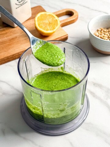 A spoon holds up vibrant vegan pesto above a food processor bowl of the same sauce. Nearby are a halved lemon on a wooden board, a bottle of oil, and a bowl of pine nuts on a marble countertop.