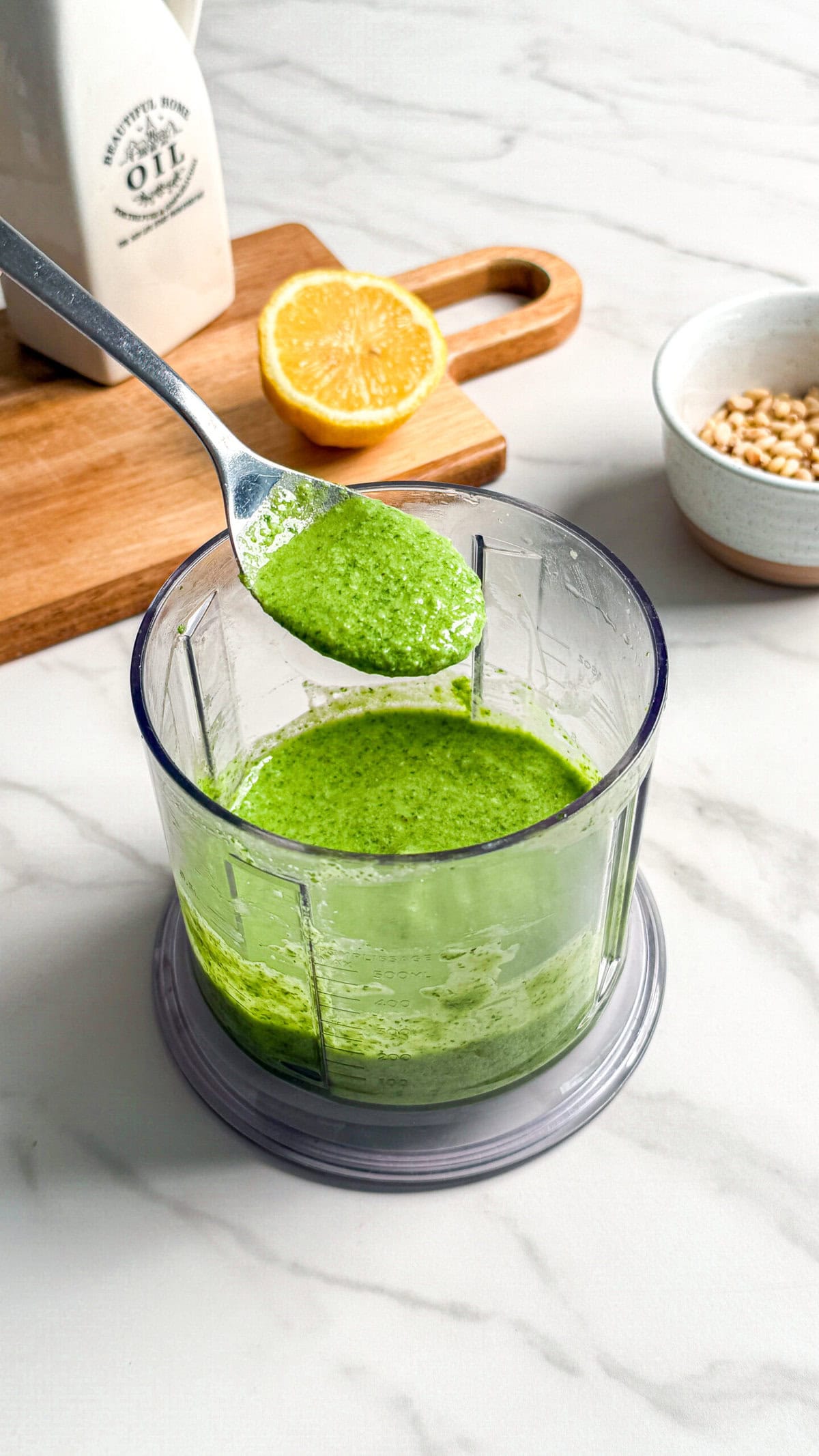 A spoon holds up vibrant vegan pesto above a food processor bowl of the same sauce. Nearby are a halved lemon on a wooden board, a bottle of oil, and a bowl of pine nuts on a marble countertop.