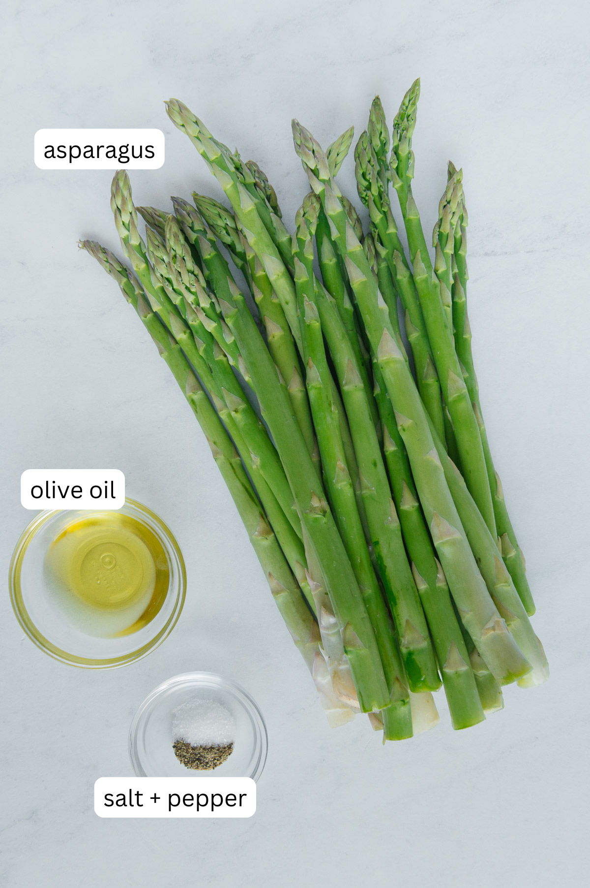 Raw asparagus next to small bowls of olive oil and seasonings with a white background.