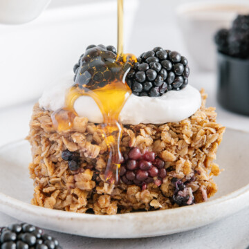 Maple syrup being drizzled over blackberry oatmeal bake topped with coconut whipped cream and two blackberries.