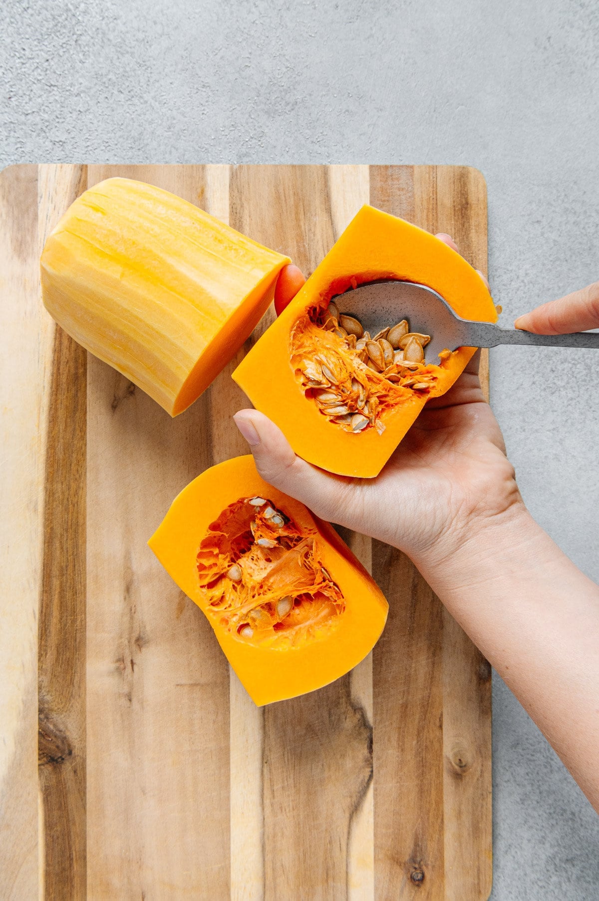 A hand using a spoon to scoop seeds out of a butternut squash over a wooden cutting board.