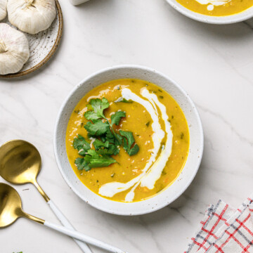 Carrot and red lentil soup in a white bowl next to fresh garlic, gold spoons and a checkered cloth with a white background