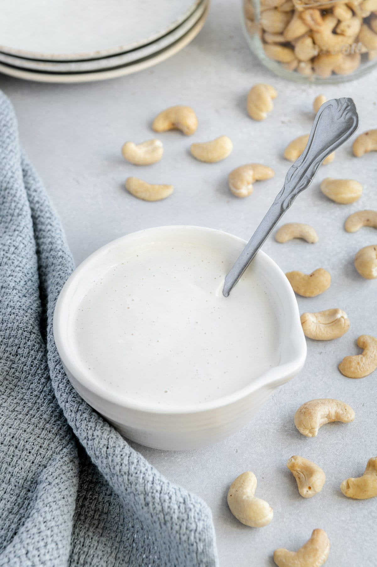 Vegan heavy cream in a white cup with a silver spoon dipped inside next to raw cashews on a light gray surface.