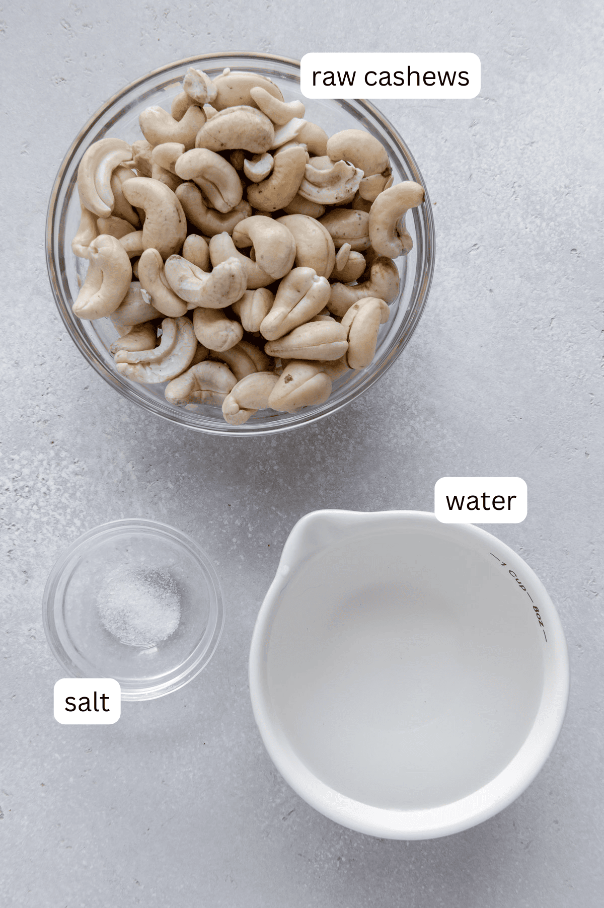 A glass bowl of cashews, a white bowl of water and a small glass bowl of salt with a gray background.
