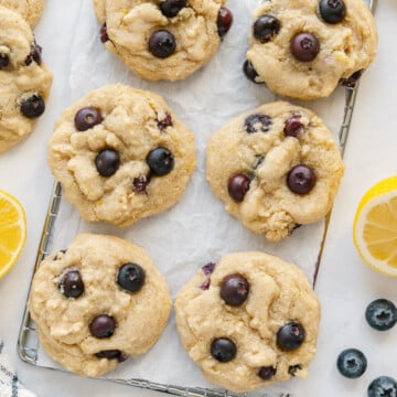 Lemon blueberry cookies on parchment paper on a wire rack next to fresh lemons and fresh blueberries with a white background.