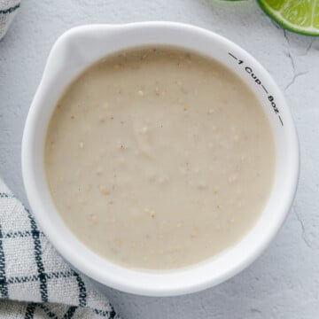 Sesame garlic lime tahini dressing in a white bowl next to sliced lime and a white checkered towel with an off-white background.