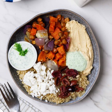 roasted sweet potatoes and onions over quinoa with hummus, vegan feta and tzatziki sauce and olives in a bowl with a white background and fork.