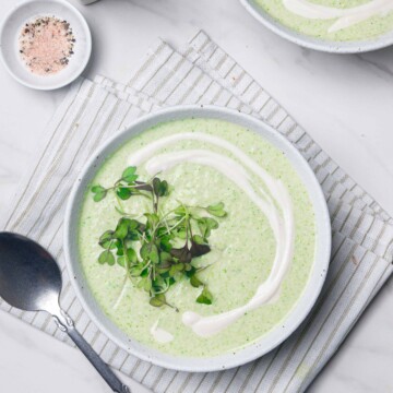 A bowl of vegan cream of broccoli soup next to a spoon on a marble white surface.