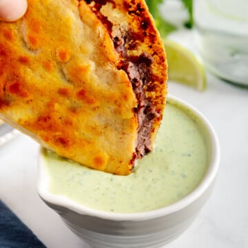 A fried black bean taco being dipped in creamy cilantro sauce.