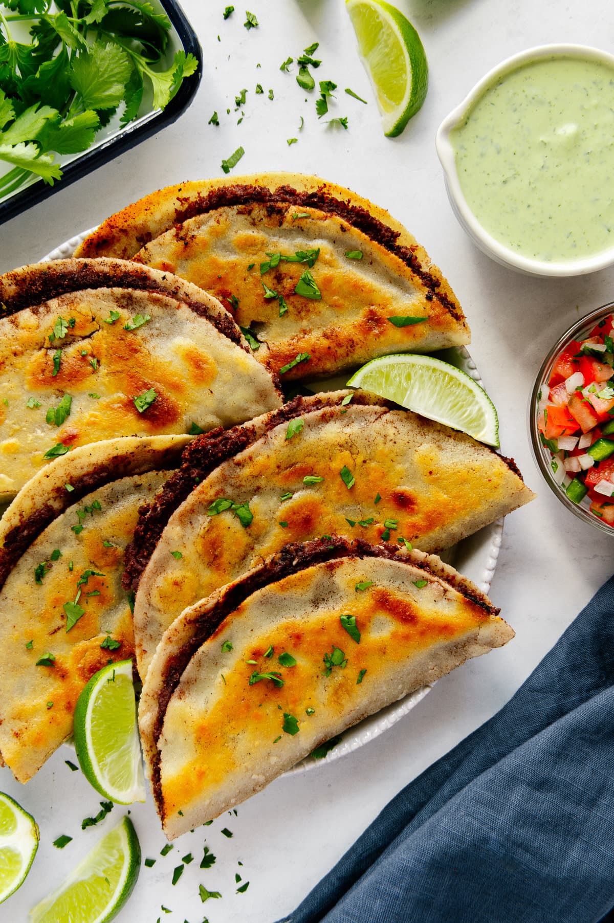 Fried black bean tacos on a white plate garnished with a lime wedge next to a bowl of salsa and cilantro sauce.