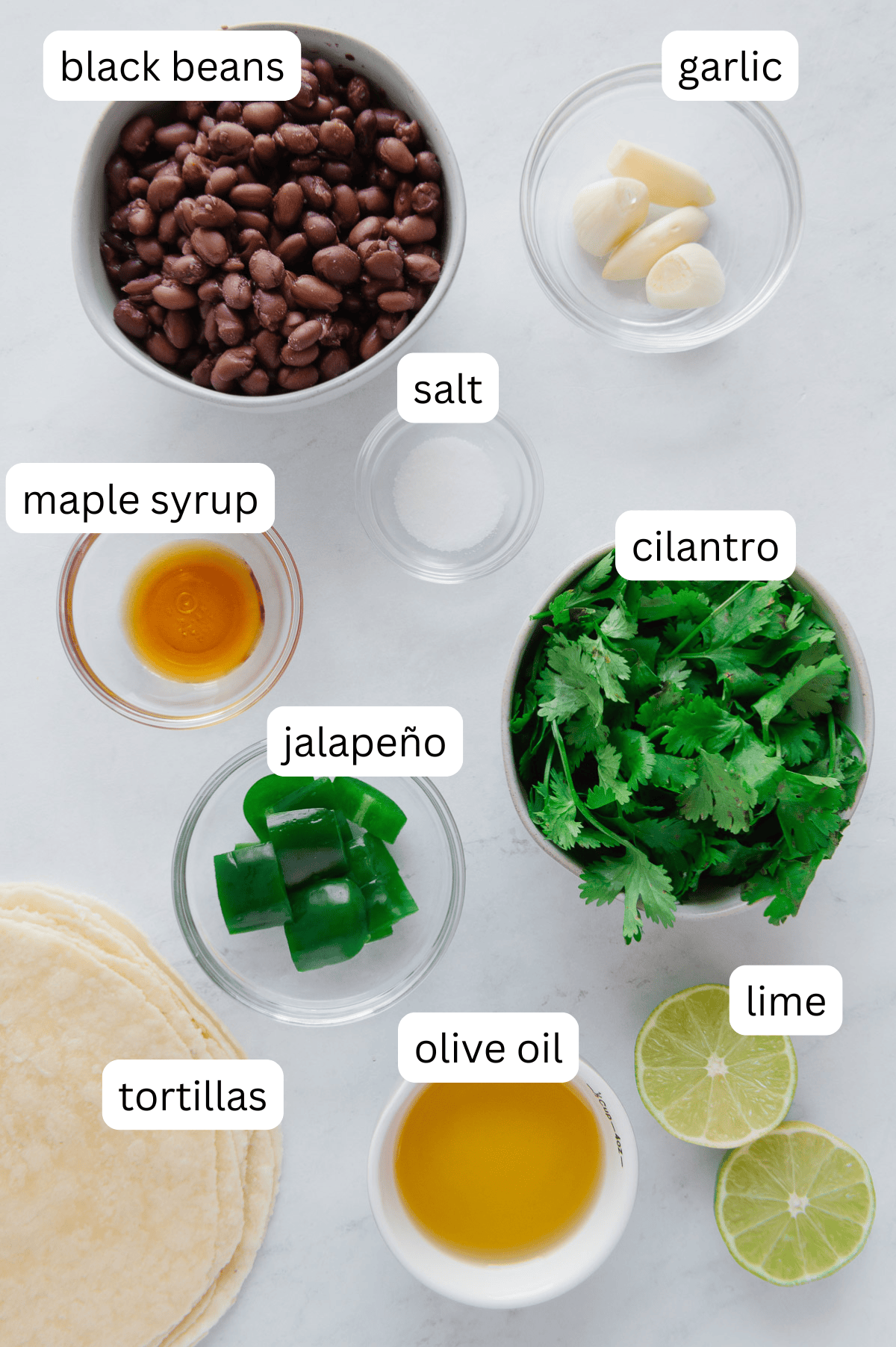 Top-down view of labeled ingredients for fried tacos on a white surface: black beans, garlic cloves, salt, maple syrup, cilantro, jalapeño slices, lime halves, olive oil, and corn tortillas.