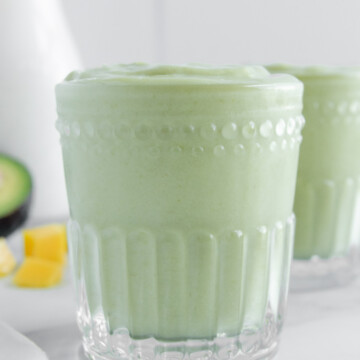 Mango avocado smoothie in a clear glass next to avocado, mango chunks and milk.