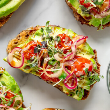 Toast topped with sliced avocado, roasted cherry tomatoes, marinated onions, tahini, date syrup and microgreens with a white background.