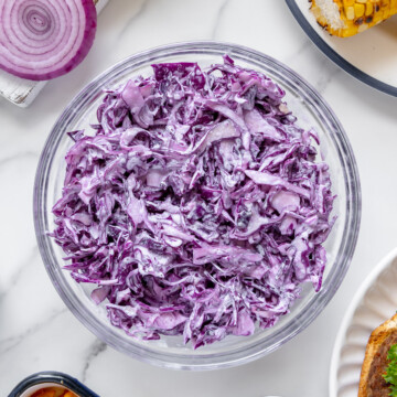 Purple cabbage slaw in a glass bowl next to other cookout foods with a white background.