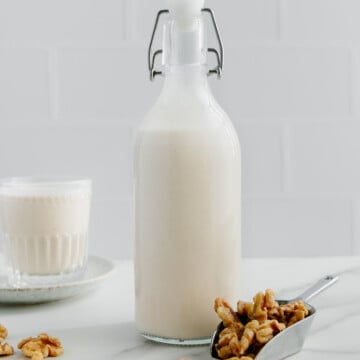 Walnut milk in a clear glass storage container with a scoop of raw walnuts beside it with a white background.