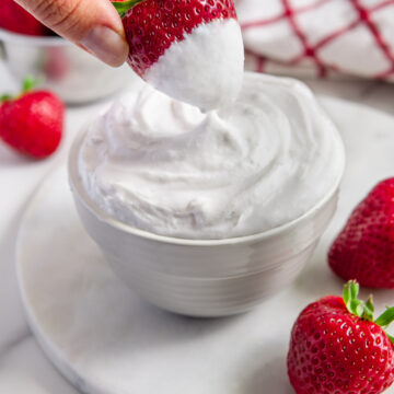 A hand dipping a strawberry into vegan whipped cream.