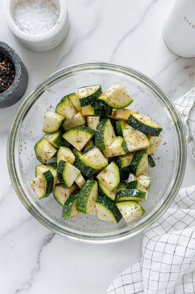 Seasoned zucchini pieces in a glass bowl with a white marble background.