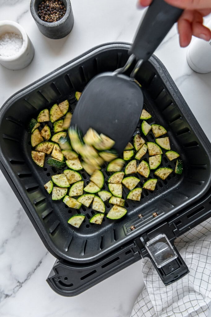 Using a black spatula to toss zucchini pieces in a black air fryer basket.