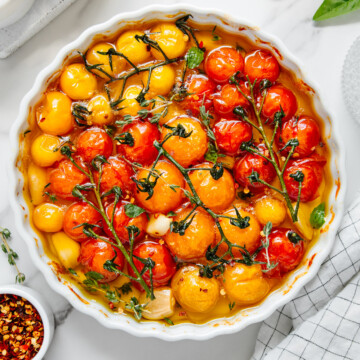 Tomato confit featuring red and orange cherry tomatoes in a white round baking dish with a white marble background.