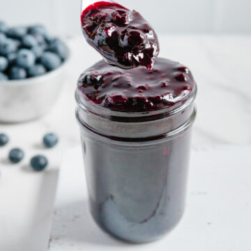 Blueberry compote in a glass jar with a spoonful lifted overtop with a white background.