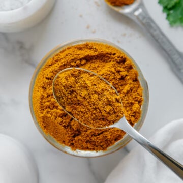 A spoonful of homemade curry powder lifted above a glass bowl of curry powder with a white marble background.