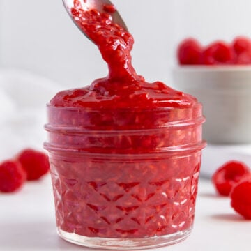 Raspberry compote in a small glass jar in front of fresh raspberries with a white background.