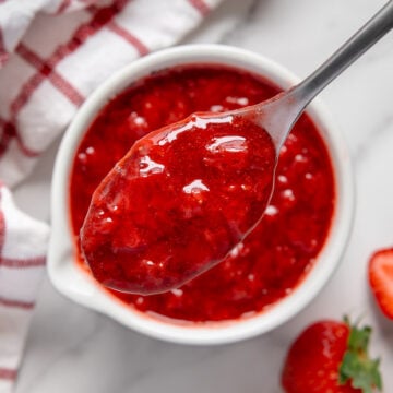 A spoonful of strawberry compote over a white bowl of strawberry compote next to fresh strawberries with a white marble background.