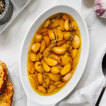 Garlic confit in a white baking dish with a white marble background.