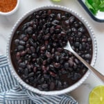 Black beans in a large white bowl served with a spoon with seasoning, fresh cilantro, sliced limes and a blue and white towel surrounding the bowl with a white background.