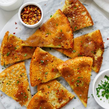 Socca wedges garnished with red pepper flakes and parsley on a round white marble cutting board.
