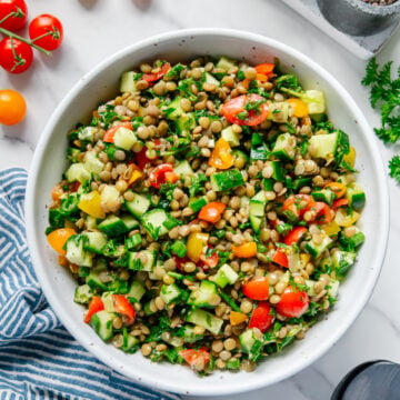 Lentil tabbouleh in a white bowl next to fresh cherry tomatoes with a white marble background.