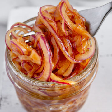 Marinated red onions in a small glass jar with a silver fork slightly lifting them with a white background.