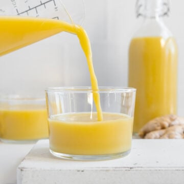 Ginger juice being poured from a glass pitcher into a small glass on a white cutting board with a white marble background.