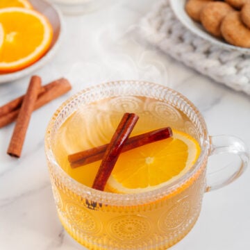 Cinnamon tea with vanilla and orange in a glass mug next to a plate of snickerdoodle cookies with a white marble background.