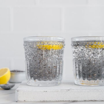 Two glasses of chia water with lemon slices floating on top on white background with lemon wedges in the background.