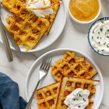 Potato waffles served on white plates next to small bowls of apple sauce and sour cream.