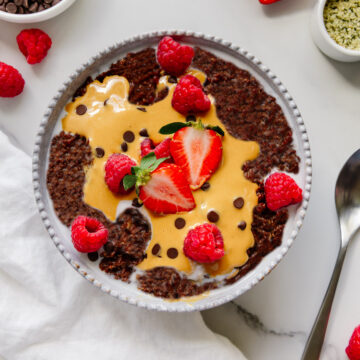 Vegan chocolate oatmeal topped with almond butter, fresh berries and chocolate chips in a white bowl with a white marble background.