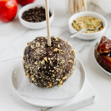 Chocolate date caramel apple topped with chopped hazelnuts on a small white plate with red apples, dates and chocolate in the background.