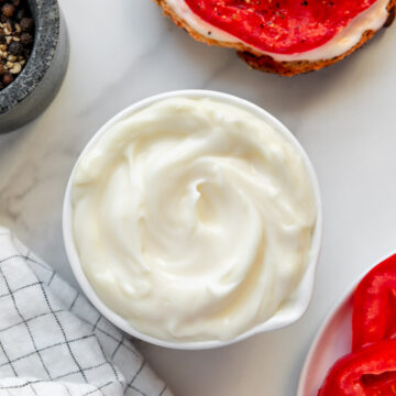 Vegan mayonnaise in a small white bowl next to an open-faced sandwich and a plate of tomatoes with a white marble background.