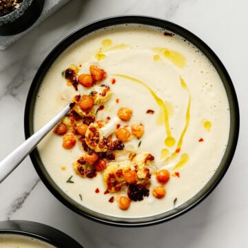 Roasted cauliflower soup in a black bowl garnished with crispy cauliflower and chickpeas with a white marble background.