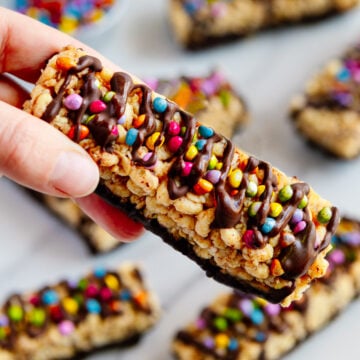 A hand holding a homemade Kudos bar with other Kudos bars out of focus on a white background.