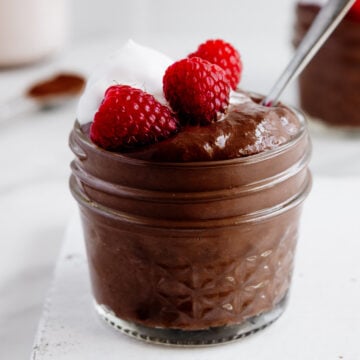 Blended chocolate chia pudding in a glass jar topped with coconut whipped cream and fresh raspberries with a white background.