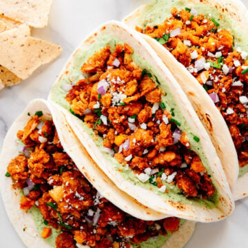 Three chickpea tacos served with diced red onion, vegan cheese and a cilantro lime sauce next to tortilla chips and lime wedges with a white marble background.