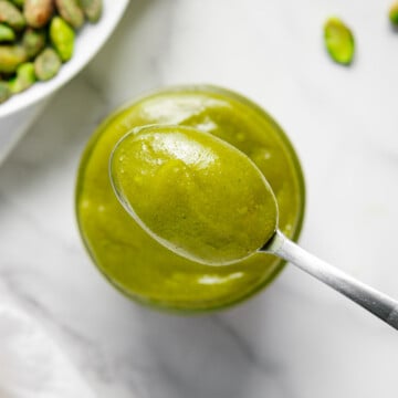 A spoonful of pistachio cream above a small jar of the cream next to a bowl of pistachios with a white marble background.