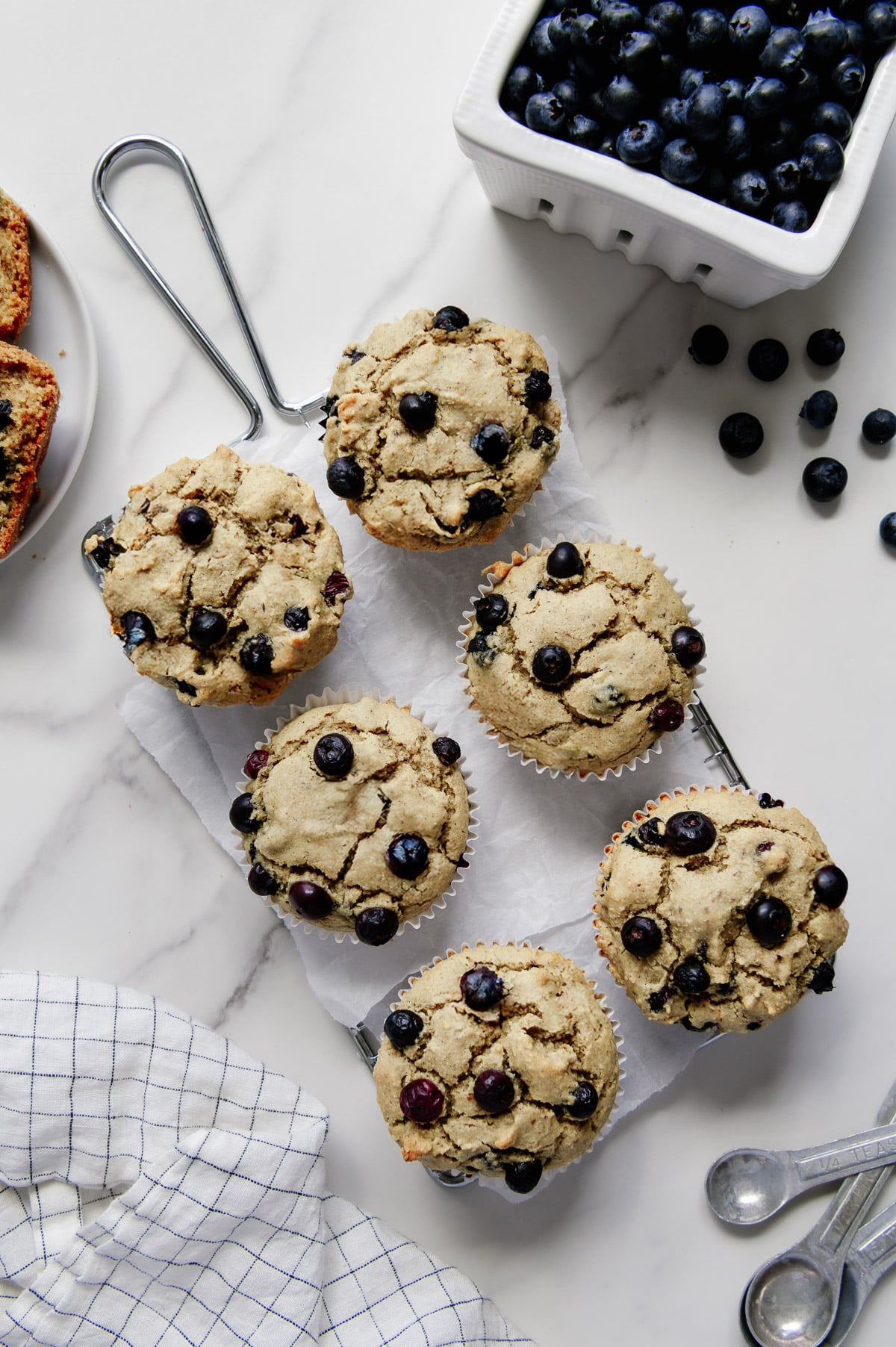 Six blueberry muffins on parchment paper on a wire cooling rack with a white marble background.